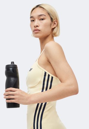 Joven mujer con cabello rubio corto y una camiseta sin mangas beige de Adidas sostiene una botella de agua negra con textura sobre un fondo liso.