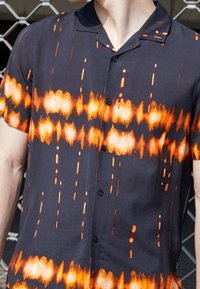Camicia a maniche corte in tessuto nero con un vivace motivo tie-dye arancione e bianco. Presenta un colletto e bottoni frontali. Texture liscia.