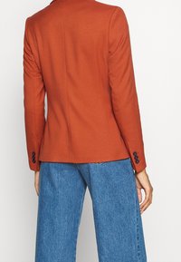 Roodbruine, op maat gemaakte blazer met gestructureerde schouders, een slanke pasvorm en vier marineblauwe knopen op elke manchette, gecombineerd met wijde denimbroeken.