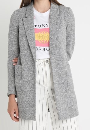 Kvinde iført grå åben-front strikket blazer, hvid T-shirt med pink og gul grafik og hvide højhåndede bukser med sorte lodrette striber.