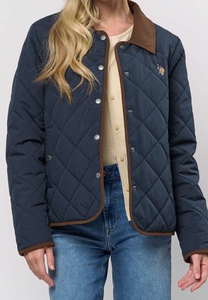 Kvinde iført en mørkeblå quiltet jakke med brune kanter og krave over en cremefarvet top og blå jeans, stående med armene afslappet ned langs siderne.