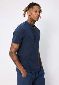Chemise henley bleu marine à manches courtes avec un col boutonné, tissu texturé et coupe décontractée. Le modèle a des tatouages et pose devant un fond uni.