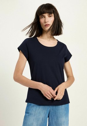 Frau mit schulterlangem dunklem Haar, die ein marineblaues kurzärmeliges Oberteil und hellblaue Jeans vor einem schlichten hellen Hintergrund trägt.