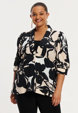 Femme souriante, portant un blazer à motifs floraux noirs et beiges avec des manches bouffantes et un pantalon noir, debout devant un fond clair uni.