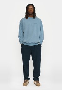 Lichtblauwe sweatshirt met ronde hals en geribbelde manchetten, gecombineerd met donkerblauwe broek en bruine sneakers met witte strepen.