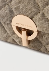 Μπεζ τσάντα χειρός από καπιτονέ ύφασμα με οβάλ κούμπωμα σε χρυσό τόνο, χαραγμένο με «VANESSA BRUNO» σε λευκό φόντο.