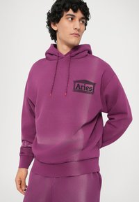 Hanorac violet cu glugă dintr-un material moale, având un buzunar frontal, glugă cu șnur și detalii cu logo-ul negru "Aries". Textură netedă.