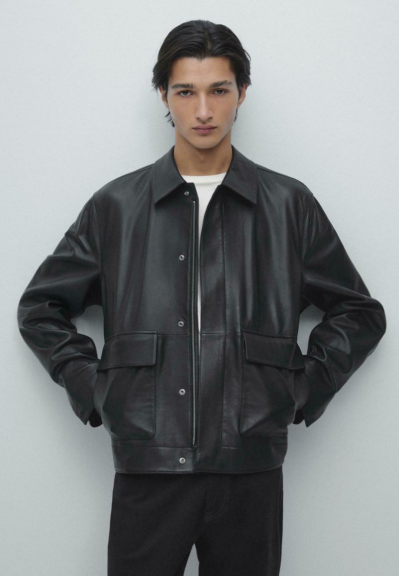 Sort læderjakke med lynlås foran, store lommer og en afslappet pasform. Har en spids krave og knapdetaljer, struktureret finish.