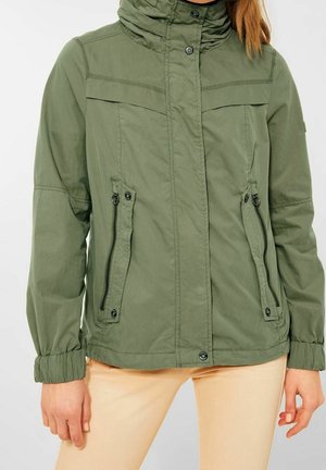 Persona con una chaqueta verde con cremallera, botones a presión y bolsillos con cremallera, combinada con pantalones beige claros.