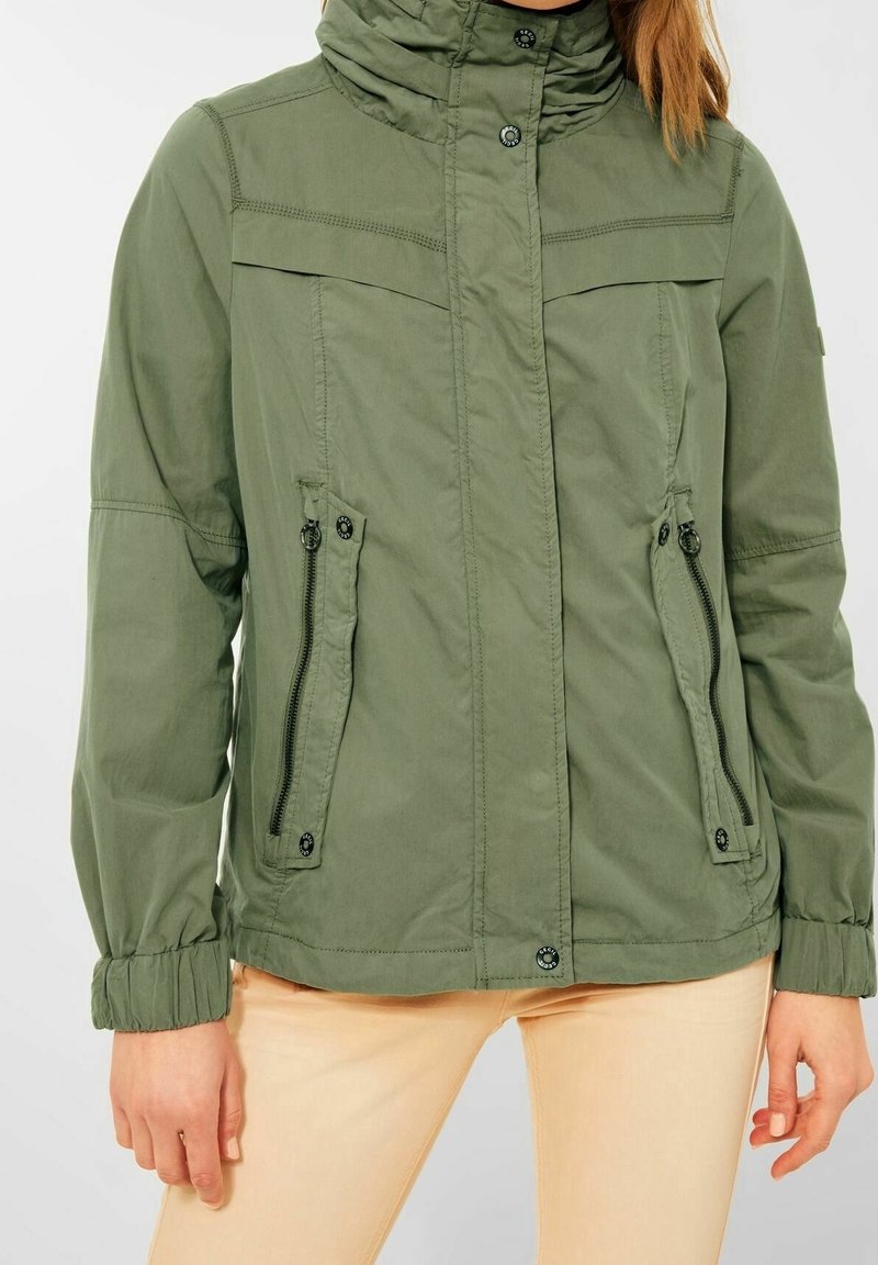 Personne portant une veste verte à fermeture éclair avec boutons-pression et poches zippées, associée à un pantalon beige clair.