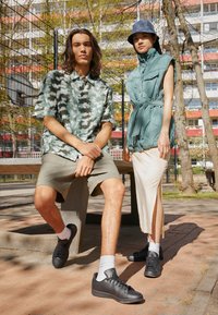 Zwei Models sitzen auf einer Bank. Eine trägt ein grünes, gemustertes Shirt und beige Shorts; die andere hat eine türkisfarbene Steppweste über einem weißen Rock an. Beide tragen schwarze Schuhe.