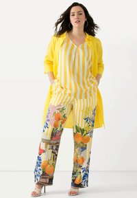 Camicia lunga gialla e leggera sopra una maglietta gialla a righe e pantaloni larghi a fantasia con disegni di frutta e fiori. Tacchi bianchi con cinturini.
