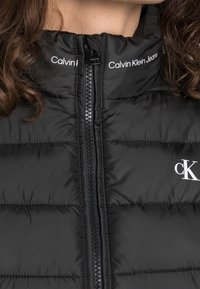 Svart puffjacka med hög krage, dragkedjestängning och horisontella quiltade sektioner. Har vit "Calvin Klein Jeans" logotyp och detaljer.