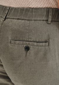 Spodnie lniane w oliwkowym kolorze z tylną kieszenią zamykaną na czarny guzik, gładką fakturą oraz zwężającym się krojem z subtelnymi paskami.