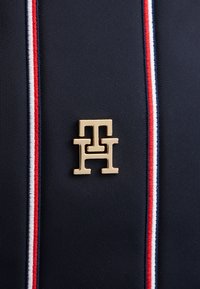 Ναυτικό ύφασμα με οριζόντια κόκκινη και λευκή ρίγα, με ένα εμφανές χρυσό μεταλλικό λογότυπο σε σχήμα αλληλοεπικαλυπτόμενων γραμμάτων "T" και "H".