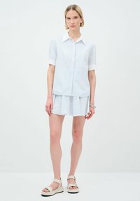 Camicia bianca a maniche corte con righe blu e colletto, abbinata a una gonna plissettata coordinata e sandali con zeppa beige.