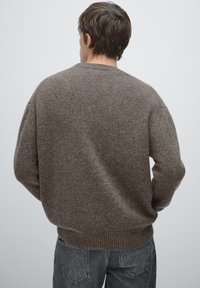 Maglione marrone a maglia con una finitura testurizzata, dotato di un collo rotondo e polsini a coste, abbinato a jeans grigi.