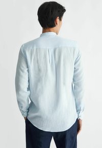 Camicia azzurro chiaro a maniche lunghe realizzata in un tessuto testurizzato, con colletto a banda, piega sulla schiena e orlo curvo. Dettagli dei polsini a bottoni visibili.