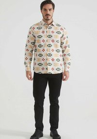 Chemise blanche à boutons avec un motif géométrique multicolore dans des tons de vert, rouge et orange ; associée à un pantalon noir slim et des chaussures noires.
