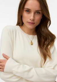 Pull en tricot couleur crème avec des manches longues et des poignets côtelés. Un collier pendentif doré avec un design complexe est porté.