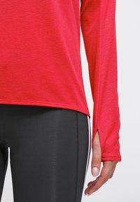 Czerwona sportowa bluzka z długim rękawem wykonana z teksturowanej tkaniny z bocznymi rozcięciami, zestawiona z czarnymi legginsami z białymi bocznymi paskami.