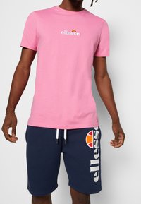 Růžové tričko s krátkým rukávem a vyšitým logem; v kombinaci s námořnickými šortkami s velkým bílým logem a oranžovým akcentem. Směs bavlny.