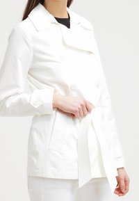 Vit dubbelknäppt jacka med dekorativa knappar och midjeband. Tillverkad av ett slätt tyg, har en klassisk krage och sidofickor.