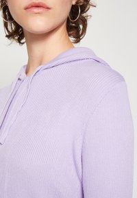 Ljuslila hoodie med ribbad textur. Har en dragsko i huvan och en avslappnad passform som framhäver tygets mjukhet och subtila lyster.