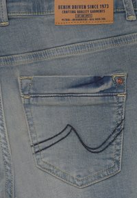 Ljusblå denimjeans har en bakficka med mörkblå sömnad i ett vågmönster, en kopparnitar och en brun etikett med varumärkesinformation.