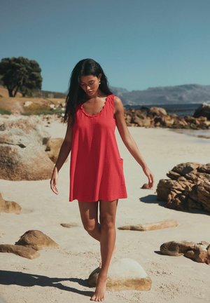 Femeie într-o rochie roșie fără mâneci care merge desculță pe o plajă nisipoasă cu stânci, munți și cer senin în fundal.