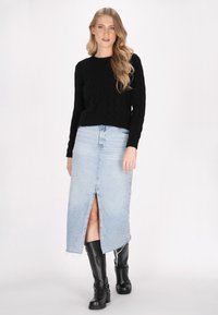 Maglione nero a treccia, gonna in denim azzurro chiaro con spacco frontale e stivali neri fino al ginocchio. L'outfit presenta un look casual ma stratificato.