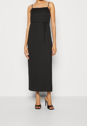 Vrouw draagt een zwarte, mouwloze midi-jurk met dunne bandjes en een taillekoord, gecombineerd met zwarte open-toe hoge hakken.