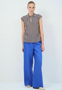 Gestreifte ärmellose Bluse in Marineblau und Beige, kombiniert mit weiten blauen Hosen. Die Bluse hat einen kragen und einen lockeren Schnitt.