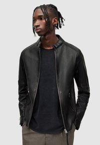 Schwarze Lederjacke mit Stehkragen, Reißverschluss und Seiten Taschen. Getragen über einem dunklen Shirt und gemusterten Hosen.
