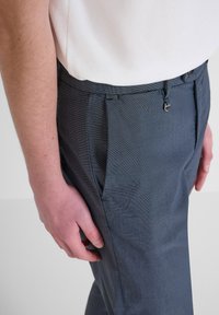 Pantalones ajustados azul marino con un tejido texturizado, que incluyen pliegues frontales, pasbelt loops y un brillo sutil. Vista lateral en primer plano.