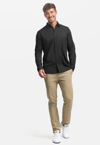 Camisa de botón de manga larga negra, chinos beige y zapatillas blancas. La camisa presenta un cuello clásico y un diseño de corte recto.