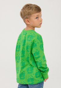 T-shirt verde a maniche lunghe con un motivo di frecce pixelate, scollo rotondo e vestibilità rilassata, abbinata a jeans di denim.