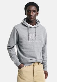 Felpa grigia realizzata in tessuto morbido, con cappuccio regolabile e logo frontale, e polsini a coste. Indossata sopra una camicia bianca e abbinata a pantaloni beige.