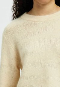 Kootud kampsun, mis on tehtud pehmest ja struktuurse tekstuuriga kreemjas kangast, horisontaalsete triipudega, mille paksus varieerub. Ümmargune kaelus ja lai lõige.