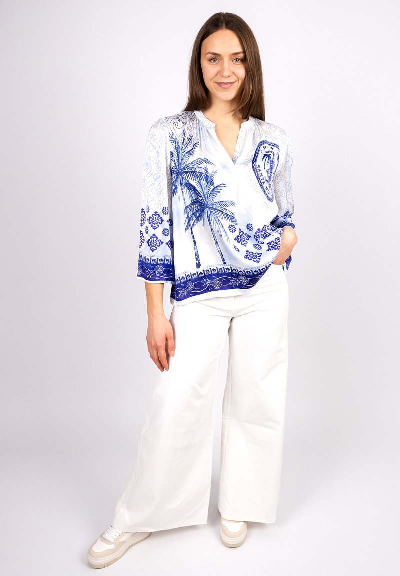 Blue and white blouse featuring a palm tree and geometric patterns, with a v-neck, long sleeves, and a relaxed fit, worn with white wide-leg pants.