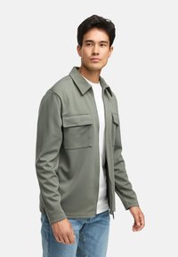 Olivgrüne Zip-up-Jacke mit zwei Fronttaschen, aus strukturiertem Stoff. Über einem weißen Hemd getragen, kombiniert mit hellblauen Jeans.