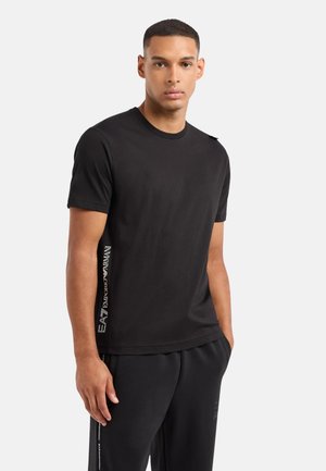Svart t-shirt med kort ärm, tillverkad av mjuk bomull, med en rund halsringning och en logotryck längs sidan. Slät textur och standardpassform.
