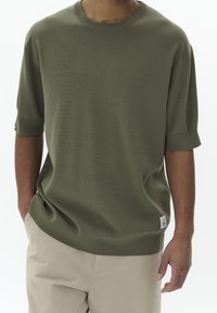 Olivově zelený svetr s krátkým rukávem, texturovaným pletením, kulatým výstřihem a malou bílou nášivkou ve spodní části. Nosené s světle béžovými kalhotami.