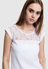 Camiseta blanca con un panel de encaje floral en los hombros. Material de algodón suave con un escote redondo y mangas cortas.