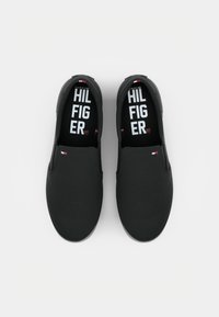 Des chaussures noires à enfiler en tissu lisse, dotées d'une semelle épaisse et de semelles graphiques affichant "TOMMY HILFIGER" en texte blanc en gras.