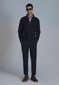 Dunkel gestreifter Cardigan mit Reißverschluss, hellblaues Hemd, maßgeschneiderte dunkle Hose und braune Wildlederschuhe. Accessoires mit schwarzen Sonnenbrillen.