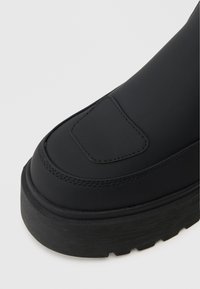 Melnas potītes zābaki ar gludu matētu virsmu, polsterētu pirkstu daļu, smalkiem šuvju akcentiem un biezu rievotu zoli. Izmanīga taisnstūra akcenta detaļa uz pirksta.