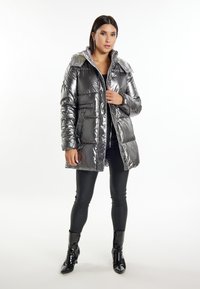 Silberner Puffer-Mantel mit glänzender Oberfläche, gestepptem Design, zwei Fronttaschen und einer faltbaren Kapuze. Trägt man über einem schwarzen Oberteil und Leggings.