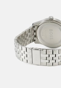 Montre en acier inoxydable argenté avec un bracelet à maillons, un boîtier rond et un cadran simple. Présente une surface texturée et une fermeture à fermoir.
