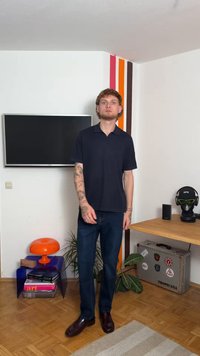 Σακάκι κοντό μανίκι σε ναυτικό χρώμα, σκούρο μπλε τζιν και καφέ παπούτσια. Τατουαρισμένος άνδρας στέκεται απέναντι σε απλή εσωτερική διακόσμηση με φυτά και τηλεόραση.